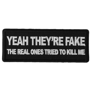 Puede incluir: Parche rectangular negro con texto bordado en blanco. La línea superior dice "YEAH THEY'RE FAKE" y la línea inferior dice "THE REAL ONES TRIED TO KILL ME". El parche tiene un borde negro.