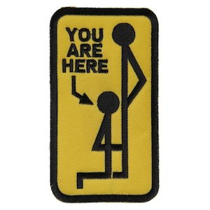 Può includere: Una toppa ricamata gialla e nera con il testo "YOU ARE HERE" e un'illustrazione di un omino stilizzato.