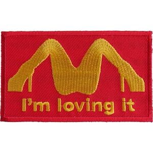 May include: A red embroidered patch with a yellow outline of a pair of legs in high heels forming the shape of the letter "M". The text "I'm loving it" is embroidered in yellow below the legs.