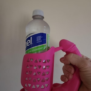 Puede incluir: Portavasos de plástico rosa con un asa con forma de mano. El portavasos tiene un diseño de malla para sujetar la botella de forma segura. La botella es transparente y tiene una etiqueta azul con el texto "Gatorade Electrolytes for your workout Zero Sugar Kiwi Strawberry KI".
