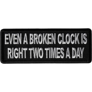 May include: Black rectangular patch with white embroidered text that reads, "EVEN A BROKEN CLOCK IS RIGHT TWO TIMES A DAY." The patch has a black border and is likely designed to be sewn or ironed onto clothing or accessories.