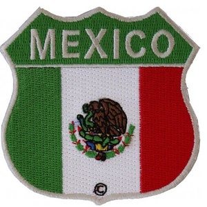 Può includere: Una toppa ricamata con la bandiera messicana a forma di segnale stradale. Il cartello recita "MEXICO" in lettere bianche su sfondo verde. La bandiera è verde, bianca e rossa con lo stemma messicano al centro.