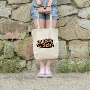 Puede incluir: Una persona sostiene una bolsa de tela blanca con el texto "Basic Witch" impreso en letras naranja y negras. La bolsa está hecha de lona y tiene un diseño simple.