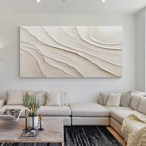 以下が含まれることがあります： モダンなリビングルームのコーナーソファの上に、白とベージュのトーンの大きな抽象画が飾られています。絵画は、波や砂丘を思わせる、テクスチャのあるレイヤーデザインが特徴です。