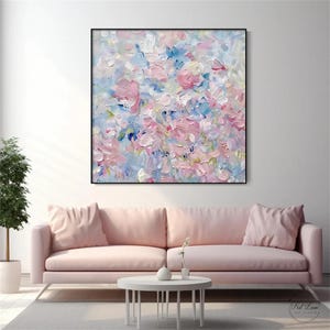 May include: A square canvas painting featuring an abstract floral composition with thick, textured impasto brushstrokes in shades of soft pink, light blue, and white. The artwork is framed in a thin black border and hangs on a plain white wall.