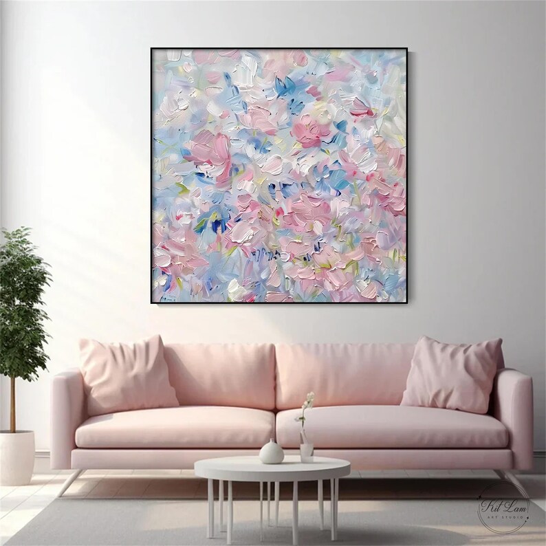 May include: A square canvas painting featuring an abstract floral composition with thick, textured impasto brushstrokes in shades of soft pink, light blue, and white. The artwork is framed in a thin black border and hangs on a plain white wall.