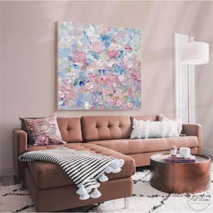 May include: A square canvas painting featuring an abstract floral composition with thick, textured impasto brushstrokes in shades of soft pink, light blue, white, and cream, displayed on a wall above a brown sectional sofa with a striped throw blanket.