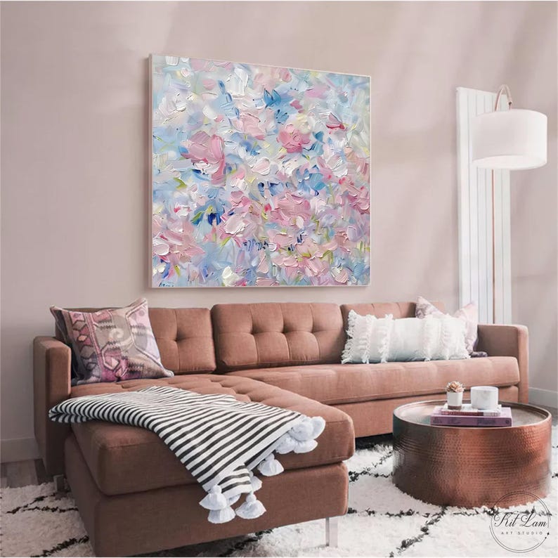 May include: A square canvas painting featuring an abstract floral composition with thick, textured impasto brushstrokes in shades of soft pink, light blue, white, and cream, displayed on a wall above a brown sectional sofa with a striped throw blanket.