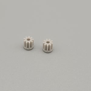 May include: Two small, light grey plastic gears with a flower-like design. Each gear has a central hole and multiple teeth around the perimeter. The gears are set against a plain, light grey background.