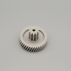 Könnte beinhalten: Ein hellgraues Kunststoffzahnrad mit zentraler Nabe und abgewinkelten Zähnen. Das Zahnrad hat eine kreisförmige Form und ist für mechanische Anwendungen konzipiert. Das Bild ist vor einem schlichten grauen Hintergrund aufgenommen.