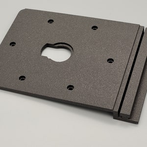 Op de afbeelding: Een rechthoekige, donkergrijze, 3D-geprinte kunststof plaat met een ronde uitsparing en meerdere gaten. De plaat is ontworpen om aan een oppervlak te worden bevestigd.