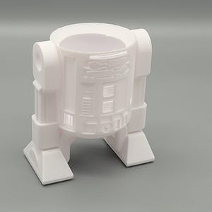 Op de afbeelding: Een witte 3D-geprinte R2-D2-vormige pennenhouder. De cilindrische behuizing heeft reliëfdetails die de panelen en kenmerken van de iconische droid nabootsen. De houder staat op een rechthoekige basis met twee voeten. Het ontwerp is een eerbetoon aan de Star Wars-franchise.