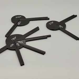 May include: Black protractors with white degree markings, including 45°, 60°, 90°, 120°, 180°, 240°, and 270°. The tools have a circular base with extending arms. The items are arranged on a light gray surface.