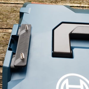 Peut inclure: Une mallette à outils bleue et noire avec une poignée noire et le mot "Sortimo" imprimé dessus. La mallette a une charnière noire et est fixée avec des vis. Le logo Bosch est visible sur la mallette.