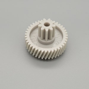 Op de afbeelding: Een grijs plastic tandwiel met een stervormig midden en de tekst "XVE" gedrukt op de zijkant.