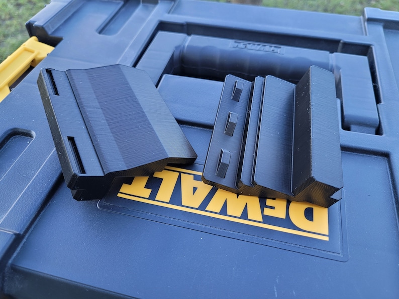 May include: Two black plastic tool organisers with a textured surface. The organisers are on top of a blue toolbox with a yellow DEWALT logo.