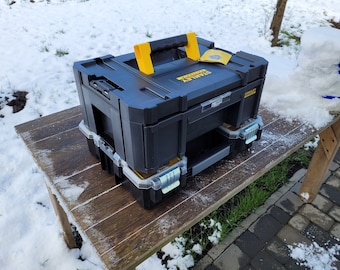 Puede incluir: Una caja de herramientas Stanley negra y amarilla con un asa en la parte superior. La caja está sobre una mesa de madera cubierta de nieve.