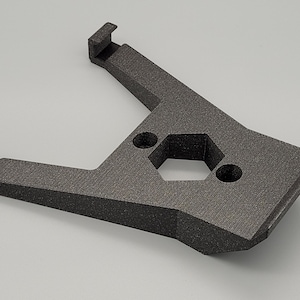 Puede incluir: Una pieza de plástico negro impresa en 3D con un agujero hexagonal y dos agujeros redondos. La pieza tiene dos brazos que se extienden desde el centro.