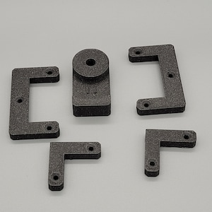 Könnte beinhalten: Fünf schwarze, 3D-gedruckte Kunststoffteile für ein Projekt. Die Teile umfassen einen quadratischen Block mit einem runden Loch, zwei L-förmige Halterungen und zwei C-förmige Halterungen.
