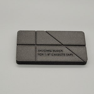 May include: A dark grey splicing block for 1/8" cassette tape. The block is rectangular with angled and straight cut lines. The text "Splicing Block for 1/8" Cassette Tape" is printed on the block.