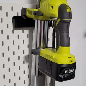 Può includere: Una pistola ingrassatrice a batteria gialla e nera, con marchio "RYOBI", è montata su una pannello forato bianco. Lo strumento include una batteria da 6,0 Ah. Un tubo flessibile si estende dal corpo della pistola.