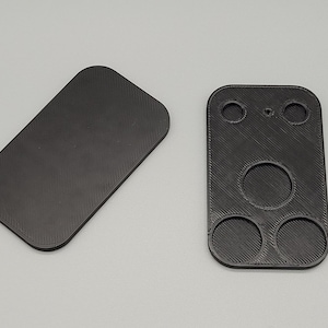 Op de afbeelding: Twee zwarte, 3D-geprinte plastic componenten. Eén is een rechthoekige plaat met afgeronde hoeken en een getextureerd oppervlak. De andere is een rechthoekig stuk met drie cirkelvormige gaten en twee kleinere gaten.