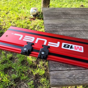 Könnte beinhalten: Rote und schwarze M18 FUEL Schienenführung für eine Kreissäge mit zwei schwarzen Knöpfen. Die Führung hat ein schwarz-weißes FUEL Logo und einen roten M18 Aufkleber.