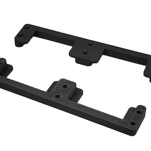 May include: Two black, rectangular brackets with rounded corners and multiple screw holes. Each bracket has a central raised section. The brackets are designed for mounting or support, likely for a mechanical or electronic device.