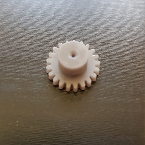 Puede incluir: Un pequeño engranaje de plástico blanco con un agujero central. El engranaje tiene una superficie lisa y redondeada y está diseñado para su uso en una máquina o dispositivo pequeño.