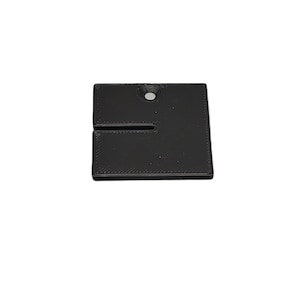 May include: A black, square-shaped product with a slot cut into the side. It has a small hole near the top. The item appears to be made of a solid, possibly plastic, material. The edges are slightly rounded.
