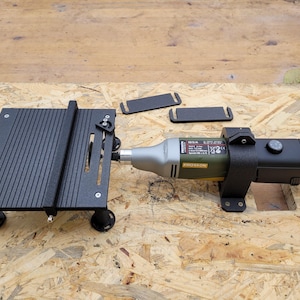 May include: A black mini table saw with a silver and green rotary tool attached. The table has a grooved surface and is supported by four black legs. Two black rectangular pieces are also present.