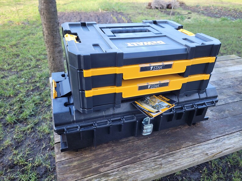 May include: A black and yellow DeWalt T-STAK tool box system with two stacked boxes and a larger black box underneath. The boxes have a yellow trim and the text "T-STAK" printed on them.