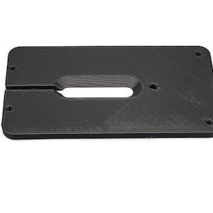 May include: A black rectangular plastic component with rounded corners. It features a central oval cutout and a slot that divides the piece. Four small holes are visible, one in each corner.