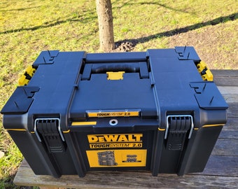 Puede incluir: Una caja de herramientas DeWalt ToughSystem 2.0 negra y amarilla con asa y cierres. La caja tiene una pegatina en la parte delantera que dice "DeWalt ToughSystem 2.0".