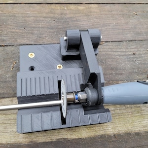 May include: A 3D printed tool for sharpening a rotary tool bit. The tool has a black base with a metal rod and a small cutting wheel. The rotary tool is grey and has a black rubber grip.
