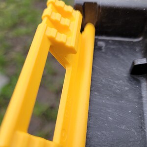 May include: A yellow plastic handle with a textured grip and a rectangular opening. The handle is attached to a black case.
