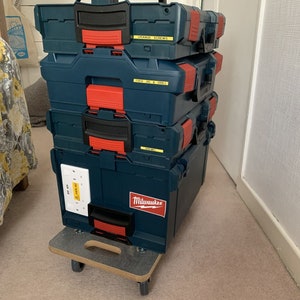 Peut inclure: Une pile de quatre boîtes à outils bleues avec des poignées rouges et des étiquettes jaunes. La boîte du haut porte une étiquette indiquant "Orange Screws". La deuxième boîte porte une étiquette indiquant "18V Drill & Driver". La troisième boîte porte une étiquette indiquant "Driver". La boîte du bas porte une étiquette indiquant "Milwaukee". Les boîtes à outils sont sur un chariot roulant en bois avec des roues.
