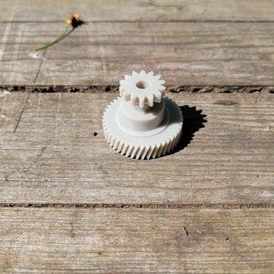 May include: A white plastic gear with a smaller gear on top. The gears are on a wooden surface.
