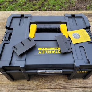Könnte beinhalten: Eine schwarze Stanley FatMax Werkzeugbox mit gelben Akzenten und einem schwarzen Griff. Die Box ist geöffnet und enthält zwei schwarze Plastikteile.