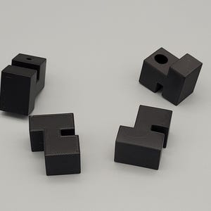 Op de afbeelding: Vier zwarte, geometrische puzzelstukken liggen op een lichtgrijze ondergrond. Elk stuk heeft een unieke vorm, met rechthoekige blokken en uitgesneden secties. De stukken lijken 3D-geprint te zijn.