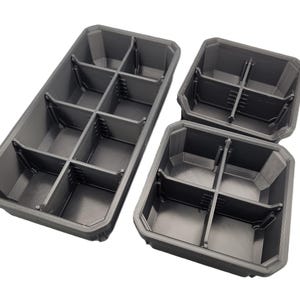 Puede incluir: Tres contenedores de almacenamiento de plástico gris con múltiples compartimentos. Los contenedores son rectangulares y cuadrados, diseñados para organizar objetos pequeños. Los compartimentos están divididos por divisores verticales y horizontales.