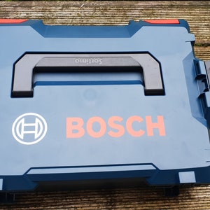 Peut inclure: Une boîte à outils en plastique bleue avec une poignée noire et un logo Bosch rouge et blanc. La boîte porte une étiquette indiquant "Softino" sur la poignée.