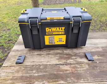Puede incluir: Una caja de herramientas DeWalt ToughSystem 2.0 negra y amarilla con una etiqueta en la parte delantera. La caja está sobre una superficie de madera.