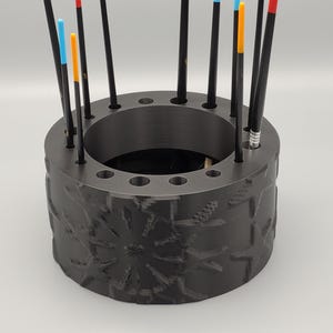 以下が含まれることがあります： 黒い円筒形のブラシホルダー。表面には花柄のテクスチャが施されています。ホルダーにはブラシ用の穴が複数あり、いくつかのペイントブラシが挿入されています。ブラシは黒い柄で、カラフルな先端が付いています。