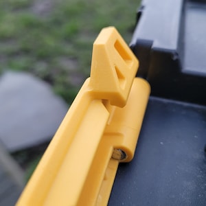 May include: Close-up of a yellow plastic latch on a black toolbox. The latch has a triangular shape with a small opening in the centre.
