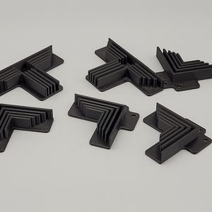 May include: A collection of six black plastic corner brackets, each with a right-angle design and multiple ribbed sections. The brackets are arranged on a light gray surface, showcasing their structural form and design.