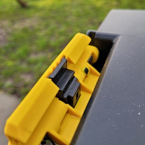 May include: Close-up of a yellow plastic latch with a black spring and a black plastic lever. The latch is attached to a black plastic surface.