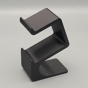 Puede incluir: Un soporte para auriculares negro en forma de Z. El soporte tiene una base plana, una sección media inclinada hacia arriba y una sección superior. Los bordes son redondeados y el soporte es de material mate.