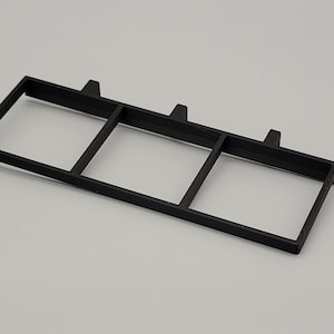 Op de afbeelding: Een zwart rechthoekig kunststof frame met drie vierkante openingen. Het frame heeft drie kleine, verhoogde lipjes langs de bovenrand. Het object staat tegen een effen, lichtgrijze achtergrond.