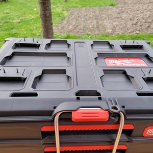 Op de afbeelding: Zwarte Milwaukee Packout opbergbox met rode accenten en een metalen handvat. De bovenkant van de box heeft meerdere compartimenten en een rood Milwaukee Packout logo.